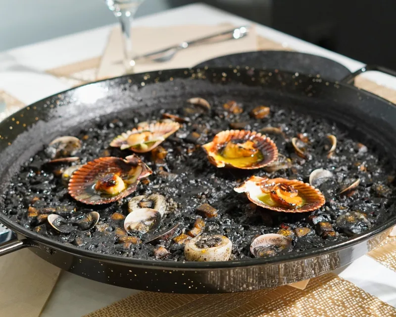 Una paella de arroz negro con zamburiñas en la mesa, junto a una copa