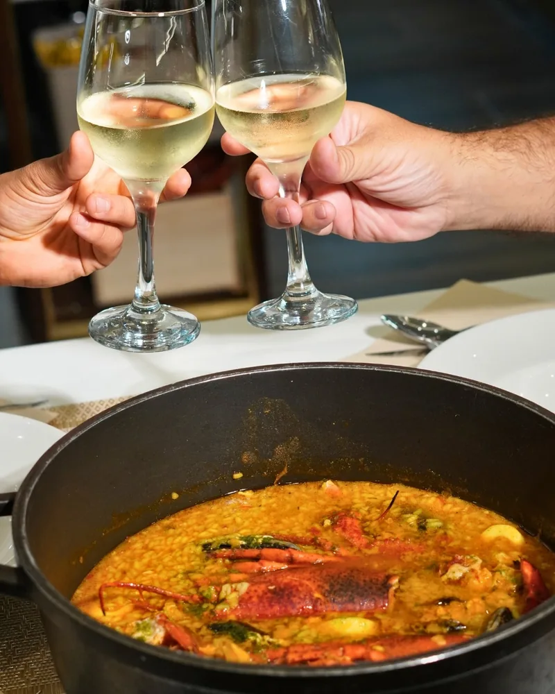 Dos copas de vino blanco brindan sobre una cazuela de arroz caldoso